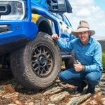 Pat Callinan knelling beside a blue wheeled drive park on top of rocks giving a thumbs up.