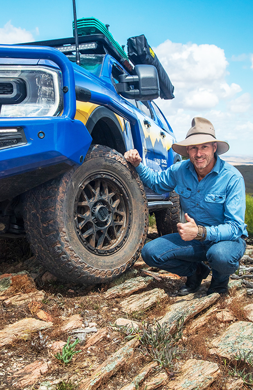 Pat Callinan knelling beside a blue wheeled drive park on top of rocks giving a thumbs up.